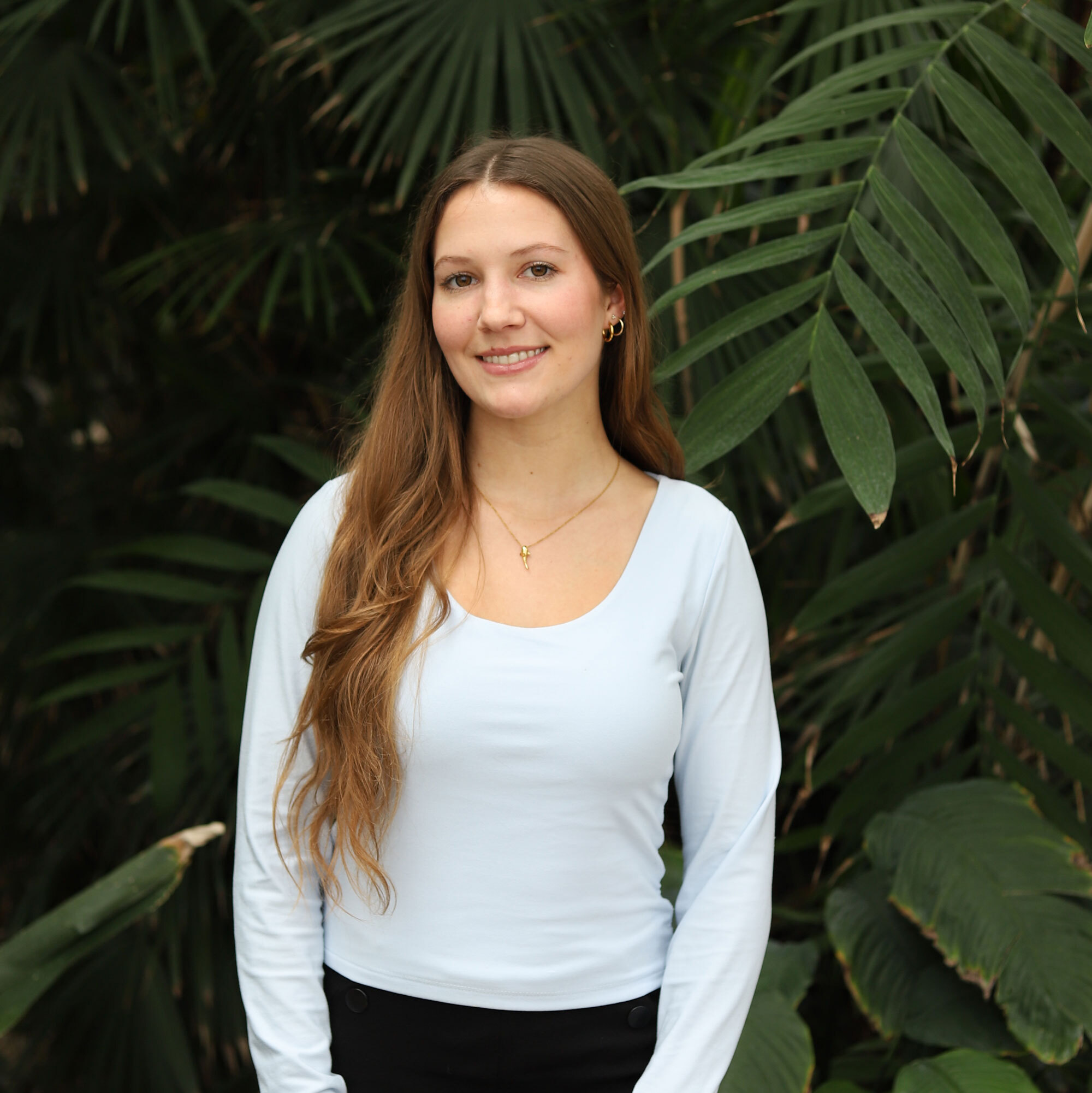 A woman with long brown hair, wearing a light blue top and gold necklace, stands and smiles in front of large green tropical leaves.